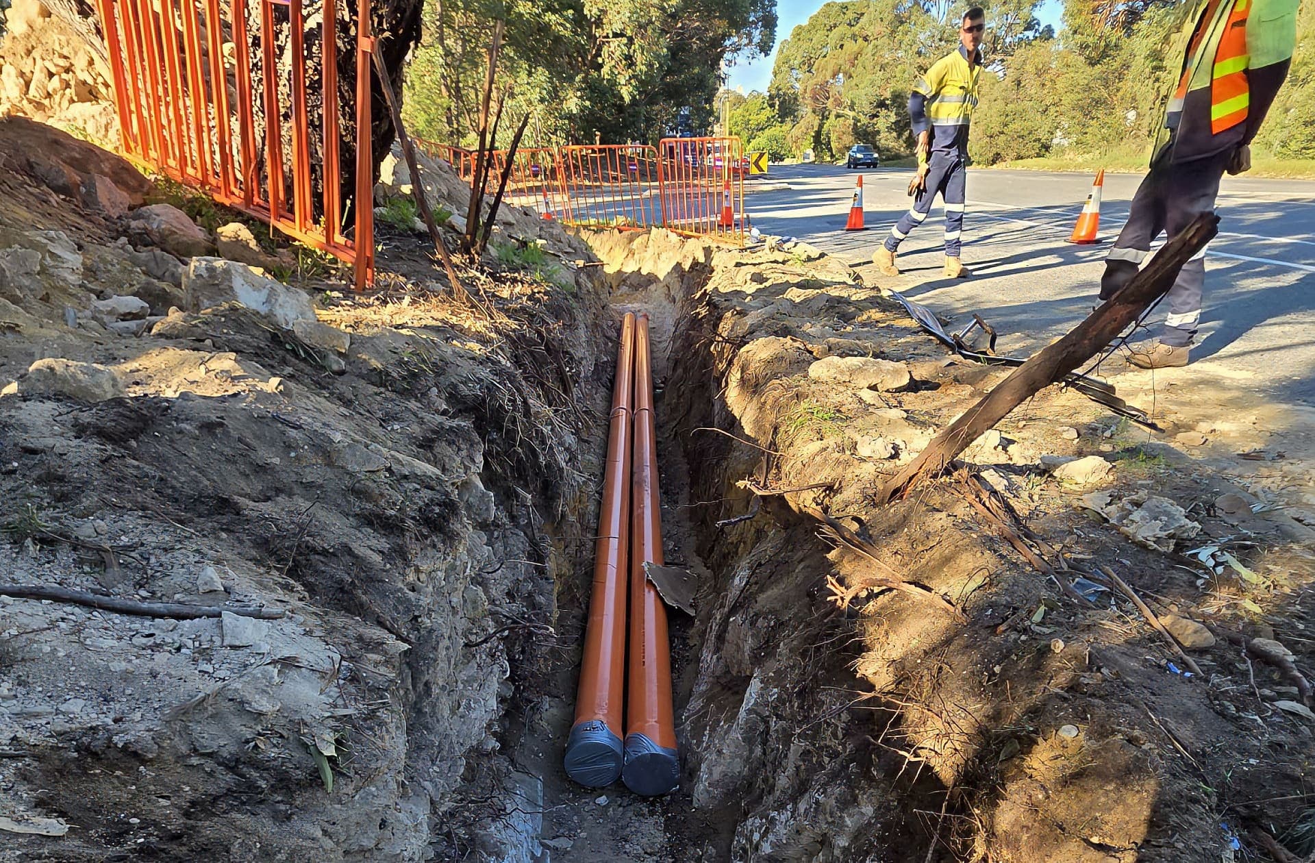 Twin orange conduits jointed in a rock trench beside Cockburn Road with the Benang crew and traffic management above