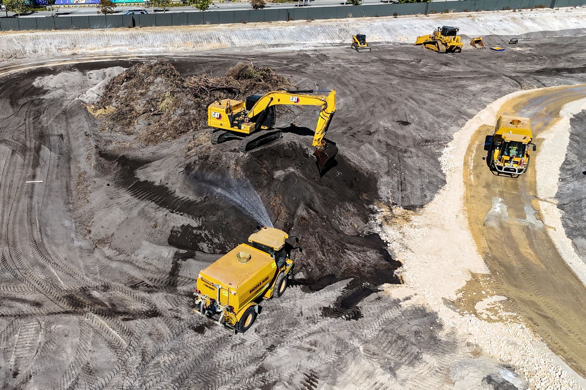 Excavator loading a topsoil stockpile while a water cart sprays for dust control on the Ellenbrook pad