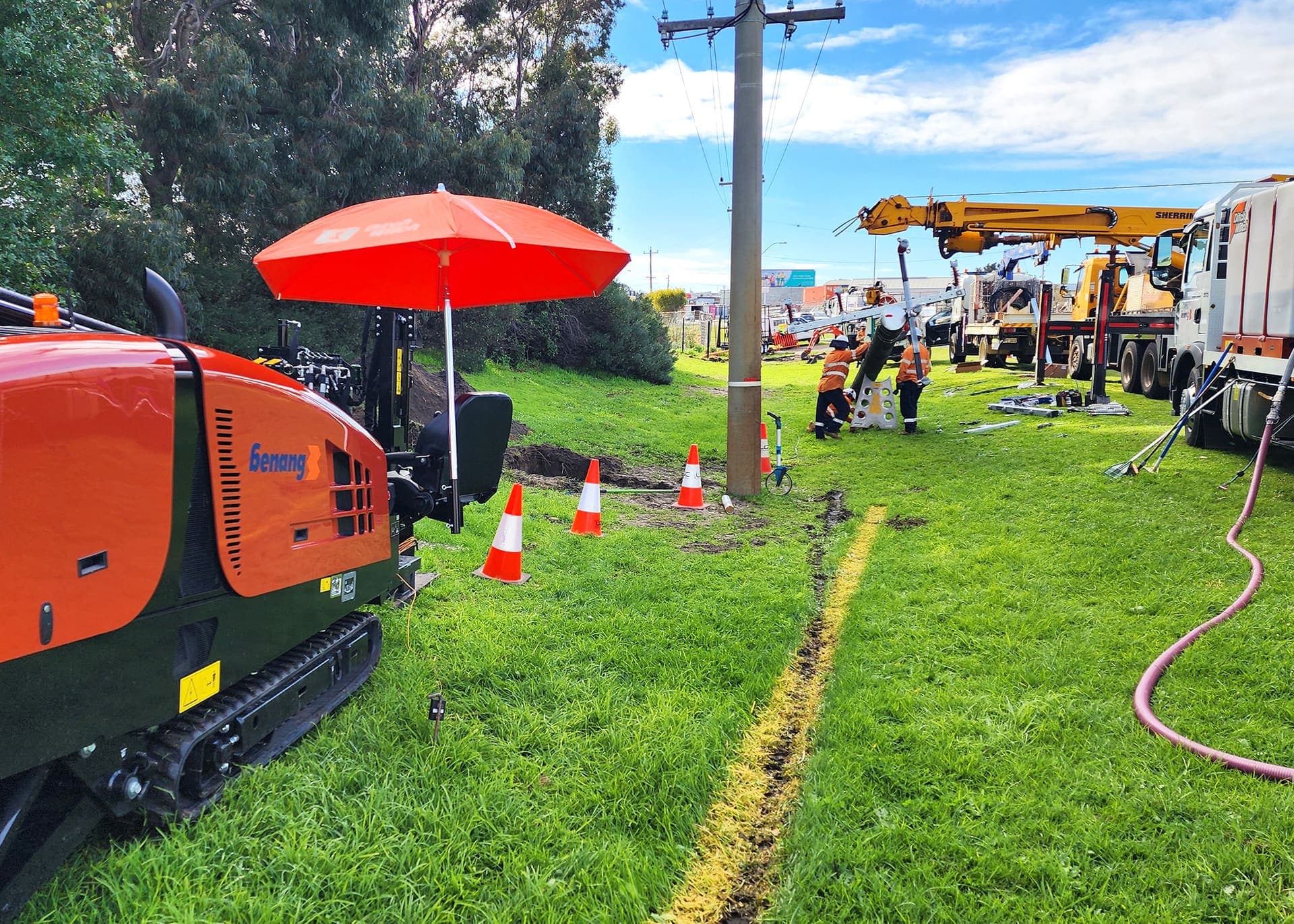 Benang HDD rig trenching along a grass verge while the crew sets a power pole in the background